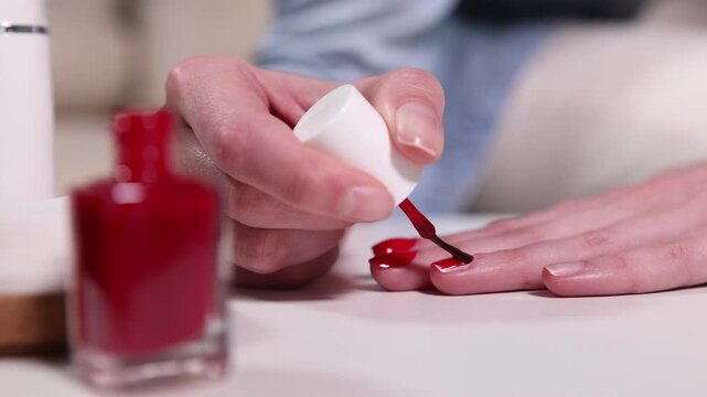 Woman applying red nail polish onto her nails at white table indoors, closeup