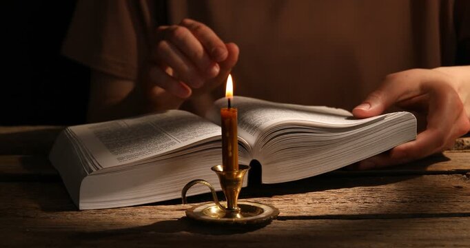 Woman reading Bible at wooden table with burning candle, closeup