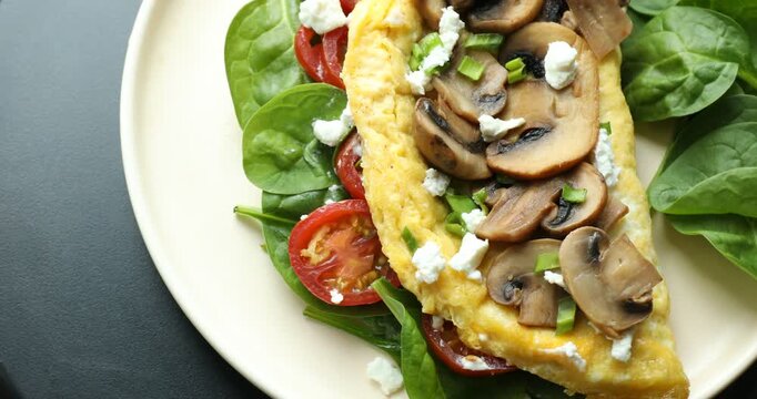 Tasty omelette with spinach, cheese, tomatoes and mushrooms on black table, spinning. Closeup view