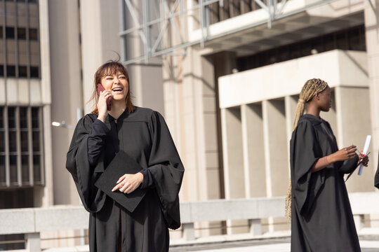 Asian and African-American women grads in black gowns laughing, holding cap and phone on terrace