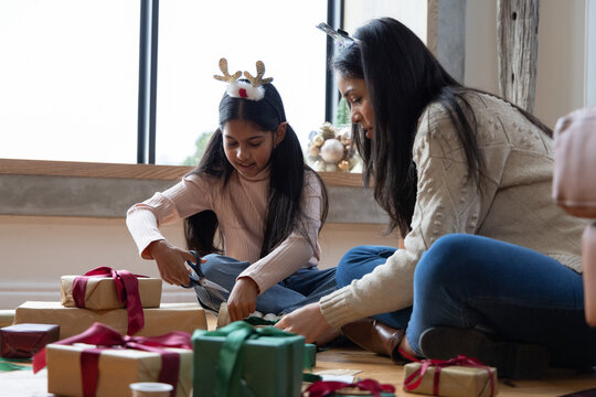 Indian mother and daughter wearing knit sweaters sitting at home wrapping kraft gifts with scissors