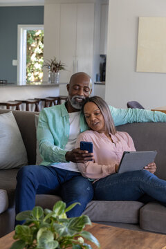 Diverse couple sitting on grey sofa at home, man showing smartphone, woman holding tablet