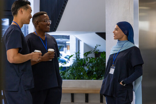 Diverse medical coworkers standing and talking in clinic lobby wearing navy scrubs holding coffee