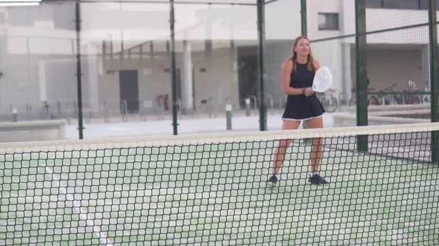 A woman with a white racket playing paddle tennis on a court next to the net,
