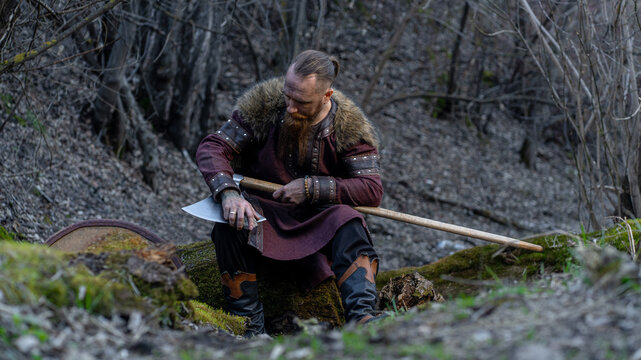 A bearded man dressed as a Viking sits sharpening an axe in a forest clearing.