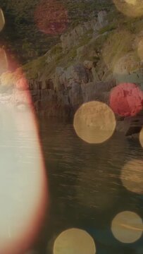 Vertical video: Warm fingertip entering left sweeping lens washing bokeh revealing rocky cove shore