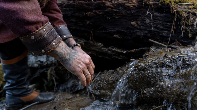 A person wearing leather bracers and boots reaches toward a flowing stream amidst a mossy, wooded landscape.