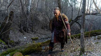 A bearded man dressed as a Viking warrior stands confidently in a wooded area, clutching a staff and appearing ready for adventure. © horimono