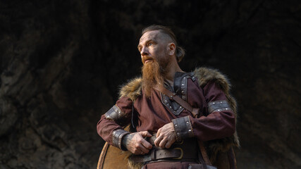 A rugged, bearded man with a contemplative gaze stands amidst a rocky landscape, clad in weathered leather and furs. © horimono