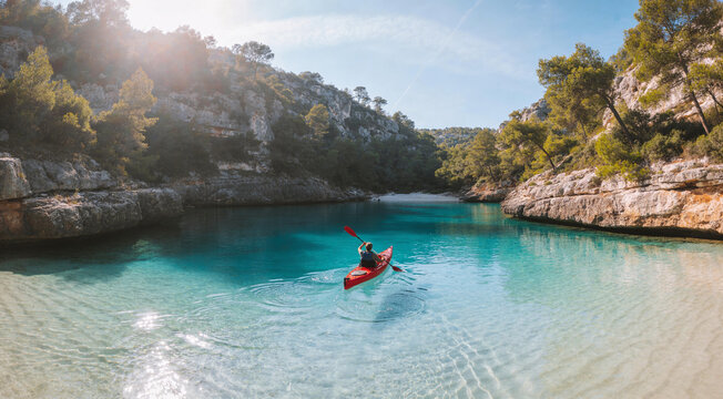 Person kayaking in a red boat through crystal-clear turquoise water in a secluded Mediterranean cove, limestone cliffs and pine trees under a bright summer sun, concept of adventure travel, coastal ex