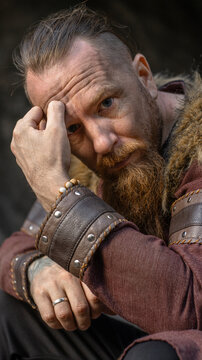 Bearded man with contemplative expression touches his forehead while wearing leather bracers and fur-trimmed cloak