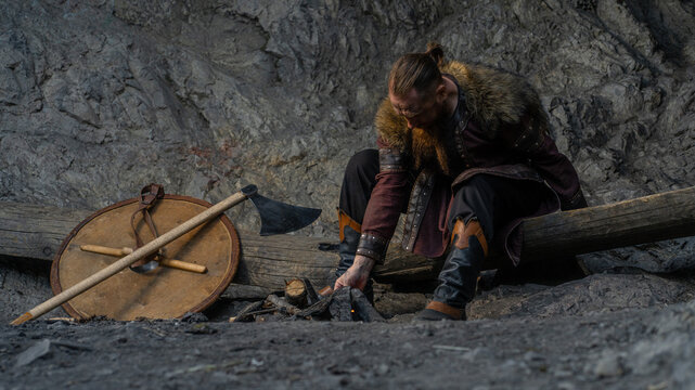 A rugged Viking warrior sits beside a campfire, sharpening his axe while resting near a shield and log.