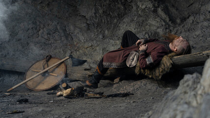 A weary Viking rests against a log, axe and shield nearby within a rocky cavern. © horimono