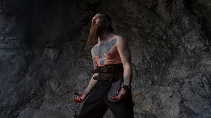 A bearded man with a tattoo on his chest stands dramatically against a rocky cave wall, gazing upwards. © horimono