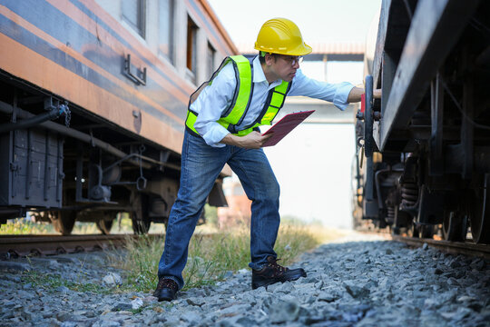 Railway engineer in safety vest and hardhat checking industrial wagon and holding clipboard at rail yard, Maintenance inspector performing safety check on train undercarriage during logistic operation