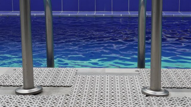 Close-up of a swimming pool entrance featuring metal ladder rails and textured poolside flooring, with clear blue water gently moving in the background