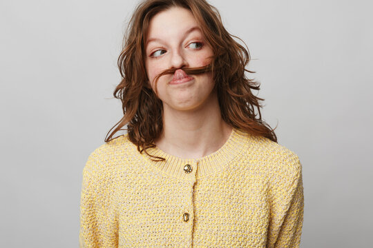 Young woman with brown hair wearing yellow sweater makes mustache with hair, looking sideways with playful expression. Studio portrait on grey background. Concept of fun and humor.