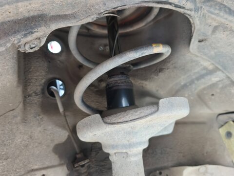 A close-up shot of an automotive suspension strut and coil spring mounted in the wheel well of a vehicle. The worn parts and surrounding chassis show the mechanical elements of the car's undercarriage