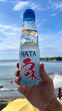 Hata Kosen Ramune marble soda Japanese beverage bottle held against a scenic coastal landscape under a bright blue summer sky - Kanagawa Prefecture, Japan, August 15, 2024