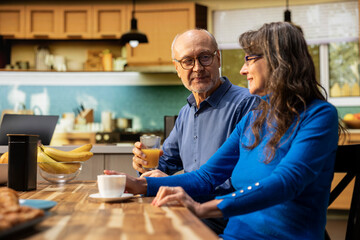 Fototapeta premium Mature couple serving breakfast together with pastry and warm coffee, bright home kitchen table. Chatting and bonding within a calm morning routine and cozy retirement lifestyle.