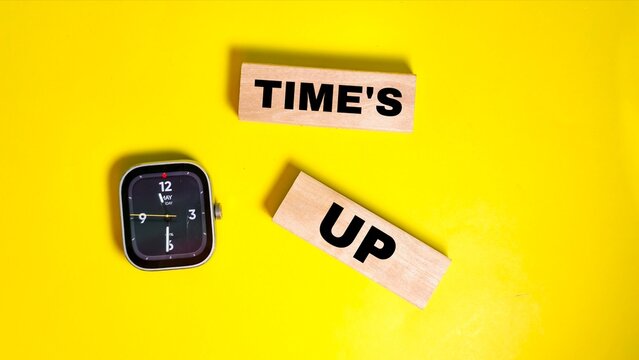 A top-down view shows a smartwatch alongside wooden blocks displaying the phrase "TIME'S UP" on a bright yellow background, representing deadlines, urgency, and the conclusion of a set period.
