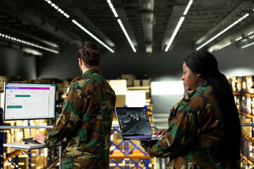 Fototapeta premium Female soldier operating digital command dashboard for aerial surveillance in a secure military warehouse. Black woman emphasizes supply chain coordination for tactical planning and defense.