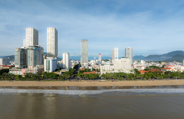 Fototapeta premium Nha Trang, Vietnam - A panoramic view of the city's coastline with modern high-rise buildings and a sandy beach dotted with people enjoying the ocean.