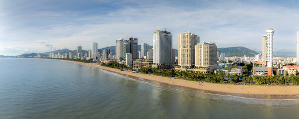 Obraz premium A panoramic view of Nha Trang beach in Vietnam, with modern high-rise hotels and buildings lining the shore. People enjoy the sandy beach and ocean waves under a partly cloudy sky.