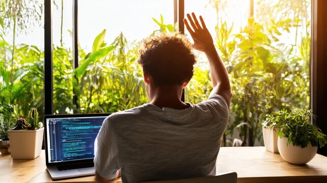 Person waves at window while coding on laptop, plants outside