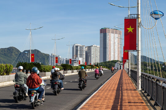 Motorcyclists ride across Tran Phu Bridge, Nha Trang,  Vietnam, on a sunny day. The bridge is decorated with Vietnamese flags and modern buildings rise in the background.