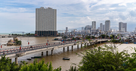 Obraz premium A scenic view of Cau Xom Bong, Nha Trang, Vietnam, with a prominent skyscraper and lush trees framing the scene. The bridge is busy with traffic, showcasing daily life.