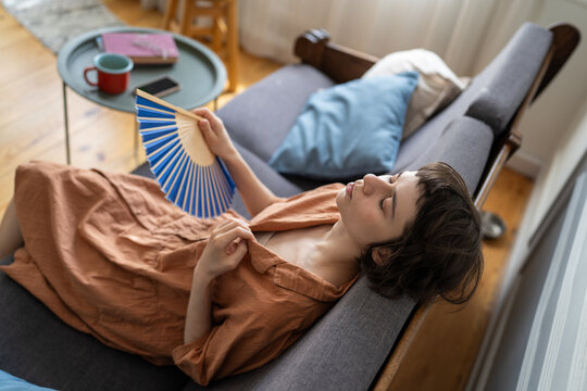Stressed woman suffering from heat at home sitting on sofa waving paper fan with closed eyes, female with dehydration and fever struggling with summer heatwave, trying to cool down with hand fan.