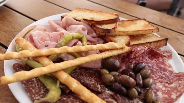Slow zoom out of italian antipasto platter with mortadella salami olives breadsticks and toasted bruschetta on wooden table outdoors.