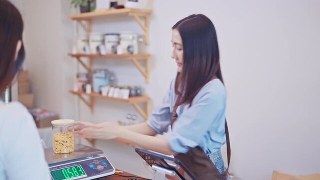 Smiling Asian female store clerk in an apron receives a glass jar of bulk food from a customer at the checkout counter of a local zero waste eco-friendly refill shop.