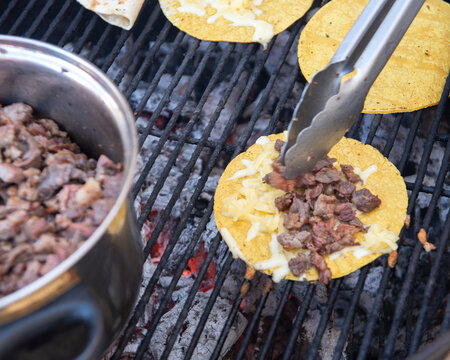 Comida en reunion familiar en el patio trasero de una casa Mexicana, con carne asada, tortillas de Maiz y harina para hacer tacos en una parrilla al carbon. Pinzas poniendo carne a una quesadilla.