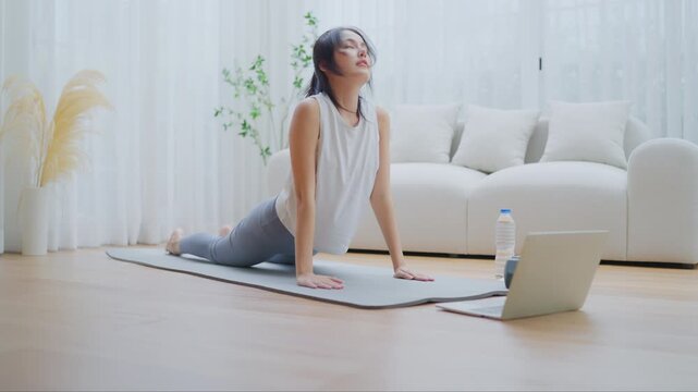 Young Asian woman stretching yoga workout on exercise mat while online training class with computer laptop in living room