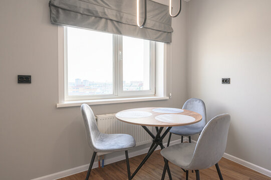 A cozy dining nook with a round wooden table, three grey upholstered chairs, large window with grey roman blind, modern pendant lights, light grey walls, wooden floor