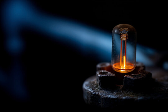 Vintage incandescent light bulb with glowing orange filament mounted on rusty aged metal socket against dark blue background showing antique electricity technology