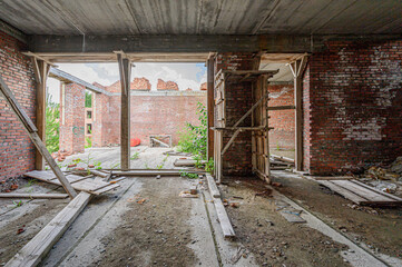 Fototapeta premium Unfinished brick building with exposed concrete ceiling, wooden scaffolding, open doorways, scattered debris, overgrown plants, cracked concrete floor, abandoned construction site ambiance.