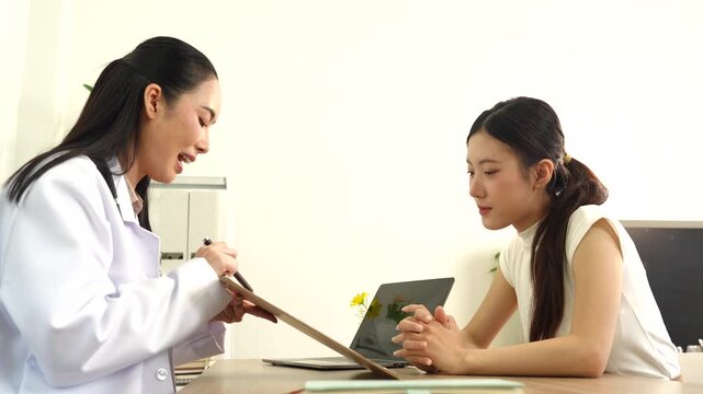 Asian female doctor consults a patient at a desk in a bright clinic, taking notes and discussing health concerns in a friendly, professional setting.