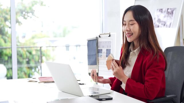 Asian Interior designer sitting in front of laptop talking with client on video call, showing architecture floor plan blueprint of residence and pantone. meeting and online conference.