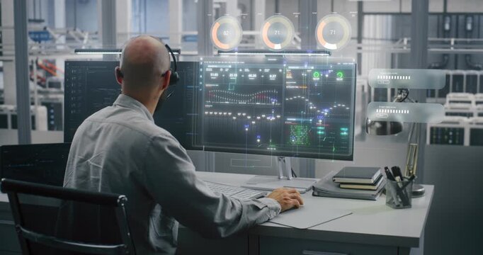 Male Engineer in Headset Sits at Desk, Typing on Keyboard and Analyzing Complex Digital VFX Data Charts, Graphs, and Percentage Holograms on Large Monitor in Futuristic Lab. Concept of Industry 4.0.