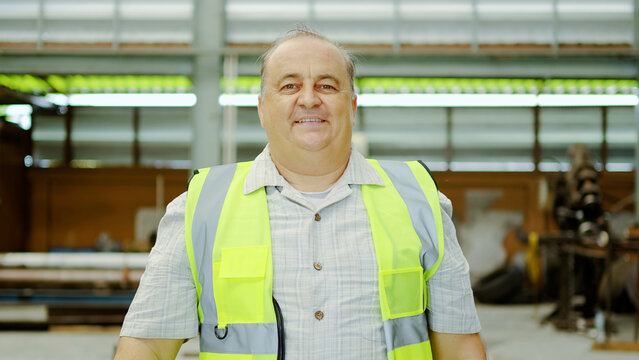 Senior Caucasian male factory worker wearing a yellow safety vest, smiling confidently while looking at the camera in a modern industrial warehouse background. Professional engineer concept photo.