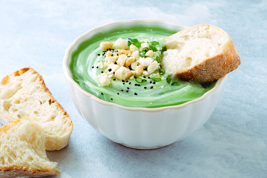 A bowl of green soup is placed on a light blue table. The soup has toppings like chopped herbs and croutons. A piece of bread is beside the bowl. It looks fresh and ready to eat. Healthy food.