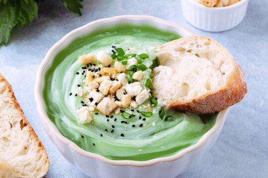 Close up of a bowl of green soup is placed on a light blue table. The soup has toppings like chopped herbs and croutons. A piece of bread is beside the bowl. Healthy food.