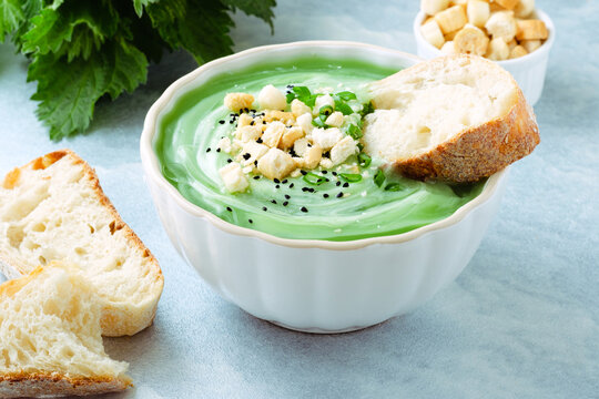 A bowl of green soup is placed on a light blue table. The soup has toppings like chopped herbs and croutons. A piece of bread is beside the bowl. It looks fresh and ready to eat. Healthy food.