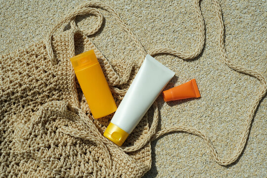 Sunscreen and lotion bottles rest on sandy ground next to a woven bag under direct sunlight. It is a warm day at the beach. Summer skincare essentials for healthy and protected skin. top view