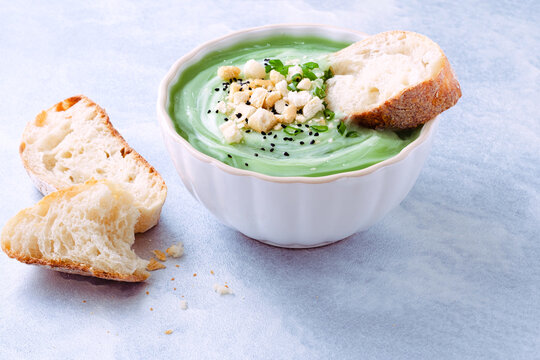 A bowl of green soup is placed on a light blue table. The soup has toppings like chopped herbs and croutons. A piece of bread is beside the bowl. It looks fresh and ready to eat. Healthy food.