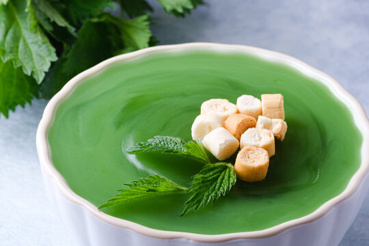 A bowl holds green soup topped with small pieces of bread. Fresh herbs are placed beside the bowl. The setting is simple, with a light surface in the background. Healthy detox food