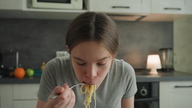 Young woman at kitchen table slurping noodles, closeup shot with steam rising, strands falling from fork, lively messy bite, warm lamp and modern cabinets in background, spontaneous informal snack
